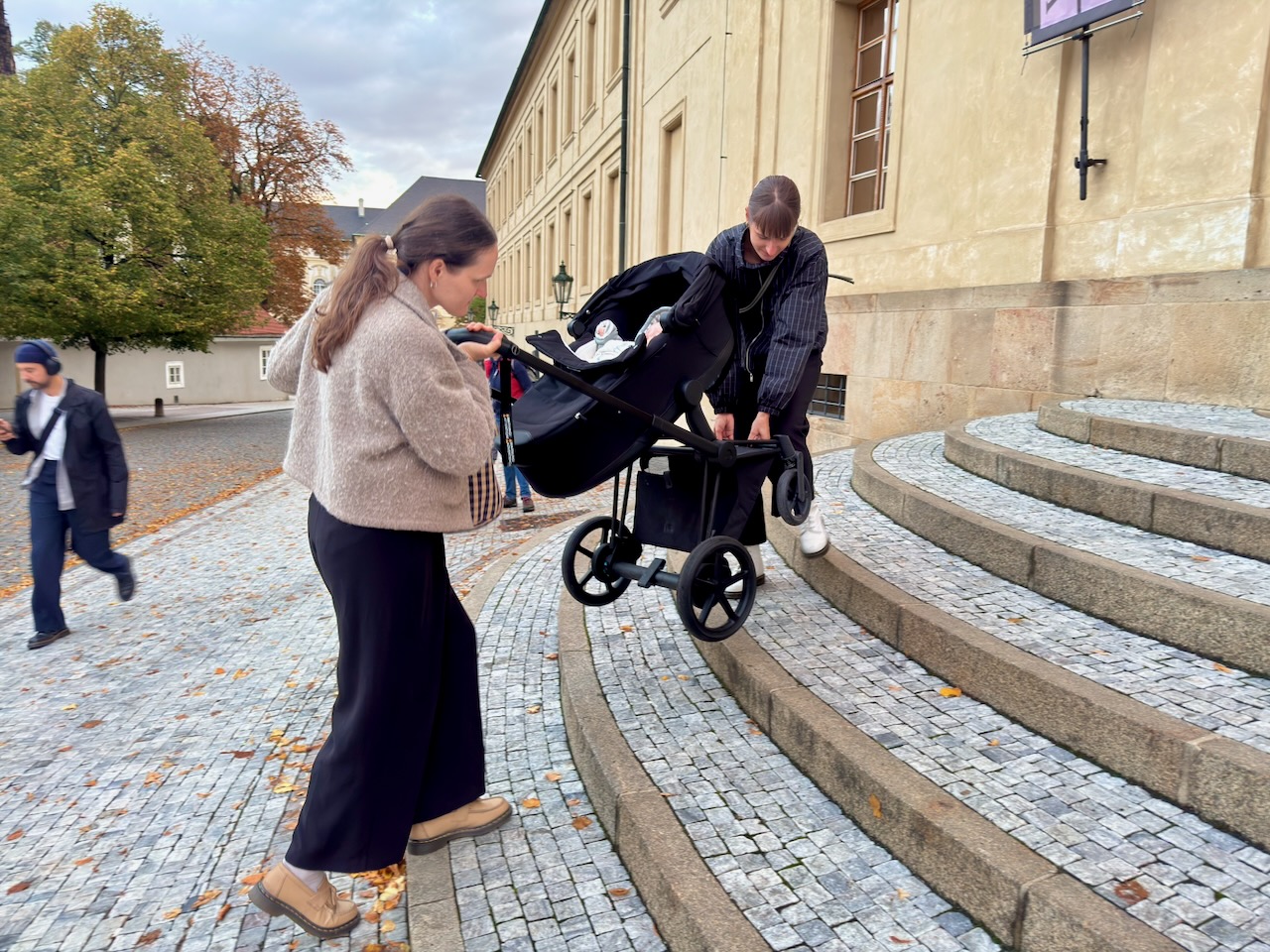 Dvě ženy vynášejí kočárek s miminkem po kamenných schodech ke vchodu do Jízdárny Pražského hradu.